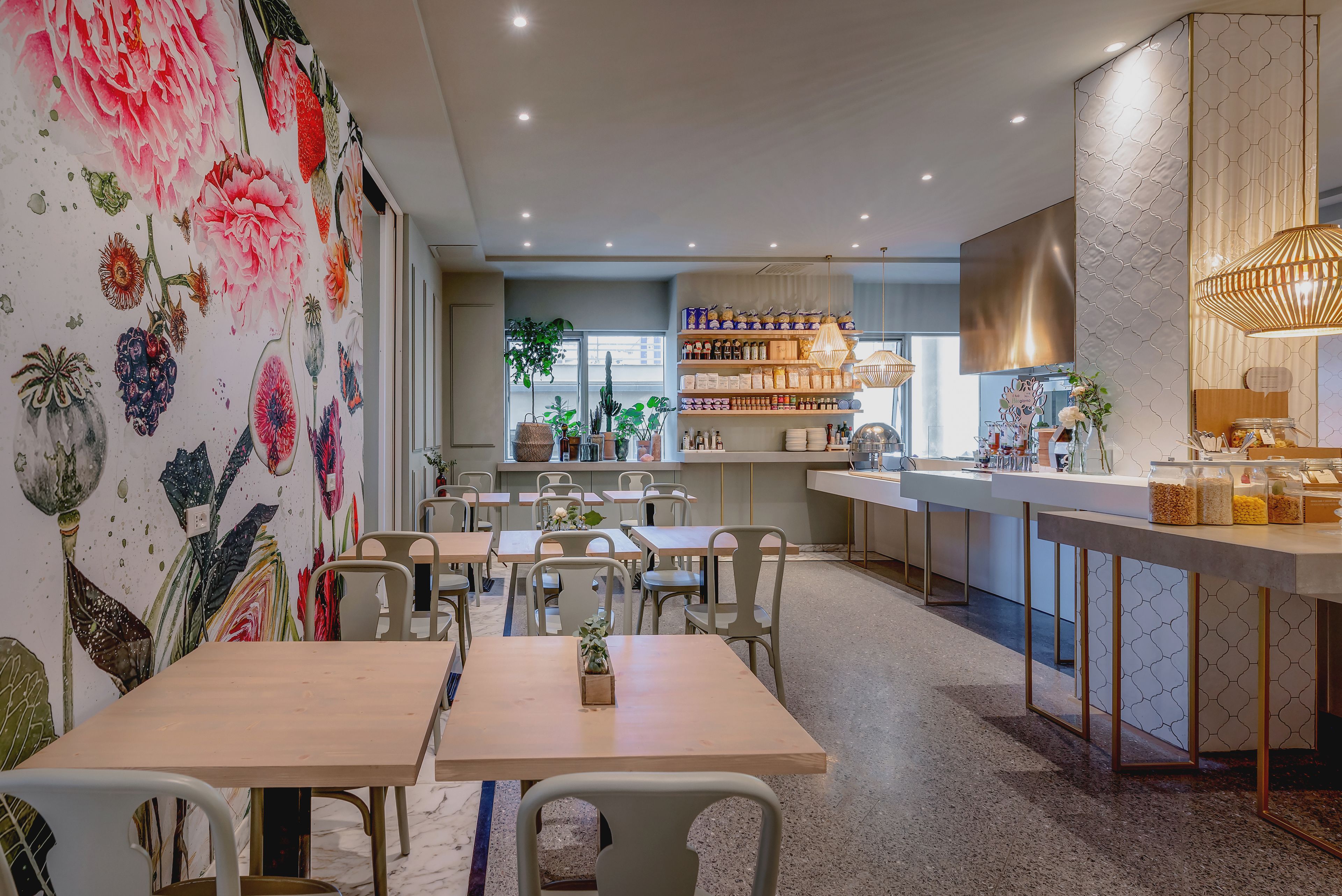 A restaurant with a floral mural, wooden tables, and a bar area with shelves of bottles and a hanging light fixture.