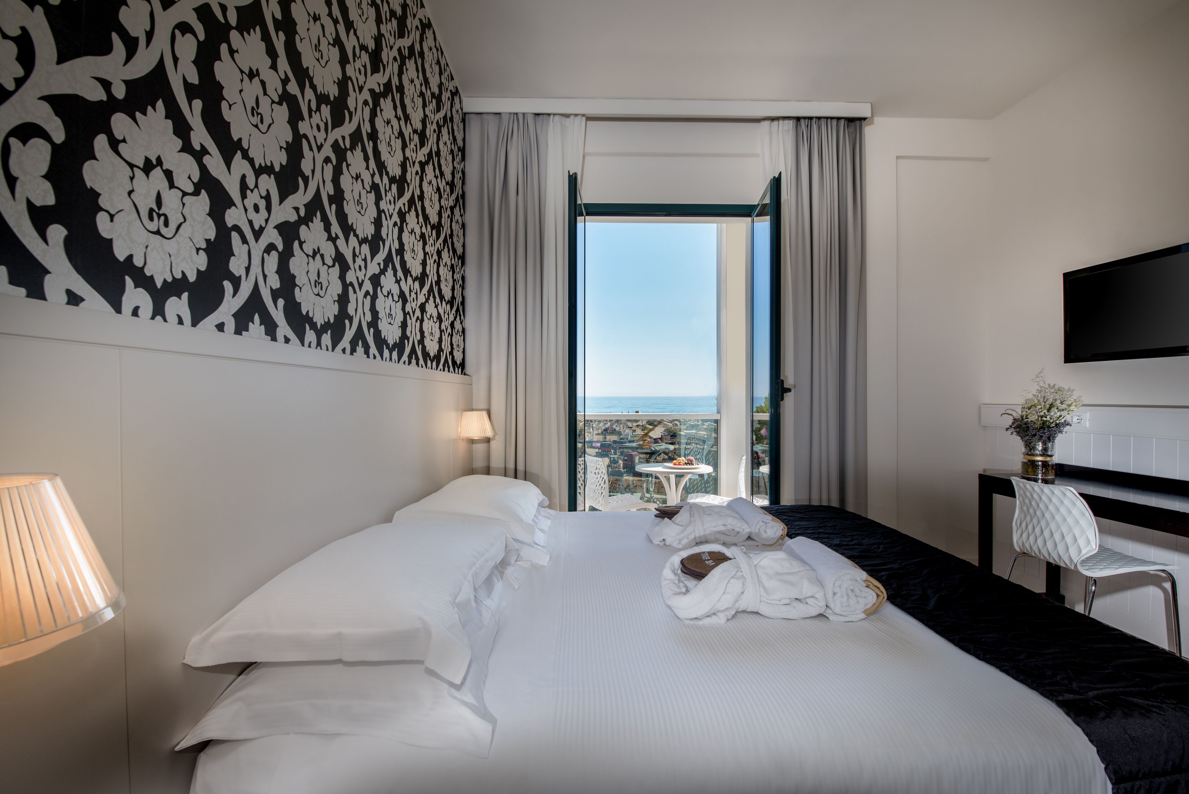 Habitación de hotel con una cama grande, televisión montada en la pared, vista a la ciudad por la ventana y una mesa con un florero.