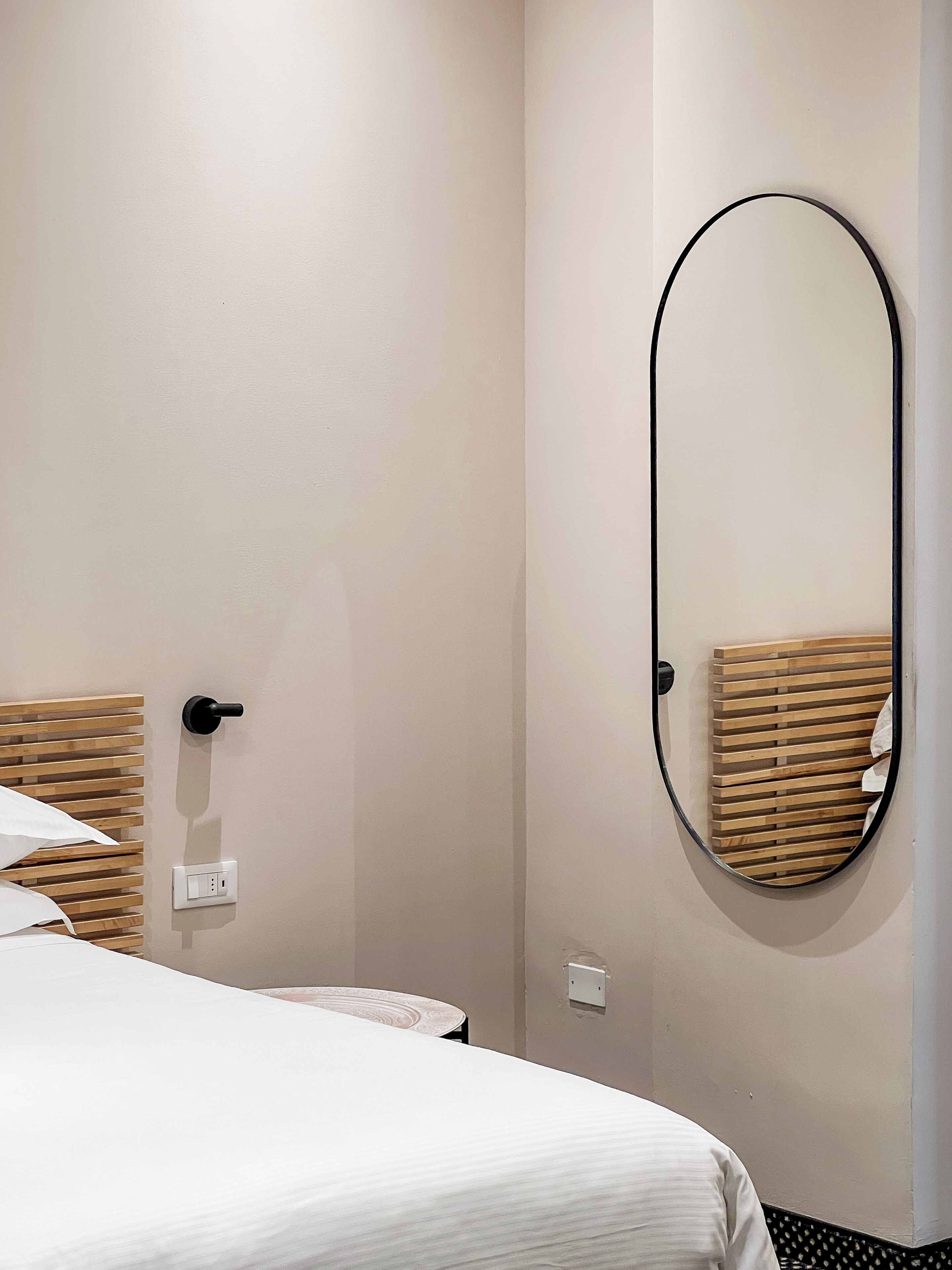 Une chambre d’hôtel avec un lit, un miroir rond, une tête de lit en bois et une prise électrique au mur.
