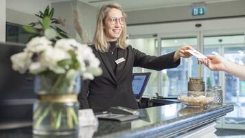 Un espace d’accueil d’hôtel comprenant un comptoir de réception avec un réceptionniste, un ordinateur, une caisse enregistreuse, un vase de fleurs et des objets décoratifs.