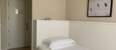 A hotel room with a single bed, a white headboard, a painting of a beach scene, and a door leading to another room.