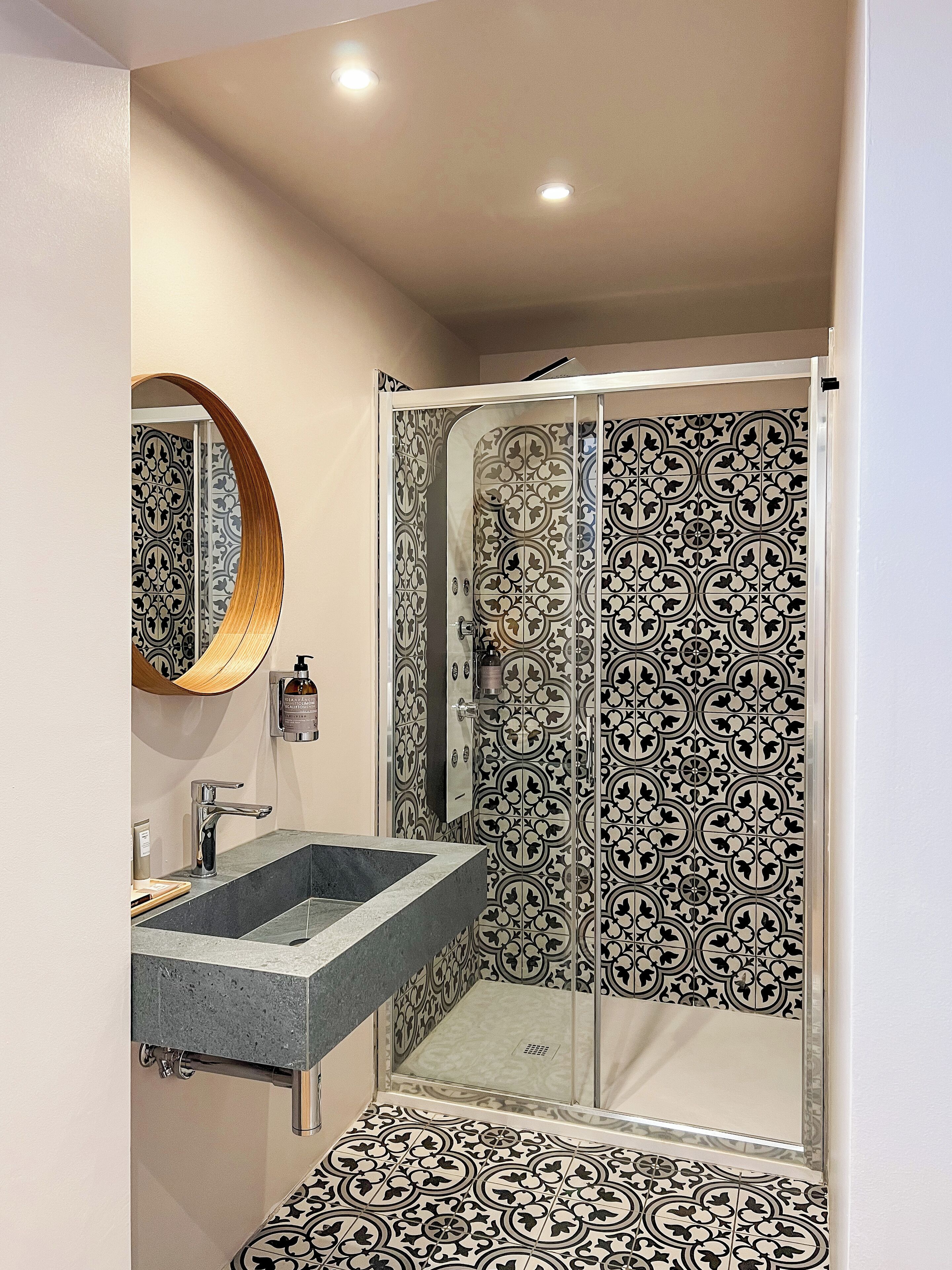 Un baño moderno con ducha envuelta en vidrio, lavabo con encimera de mármol y azulejos estampados en paredes y suelo.