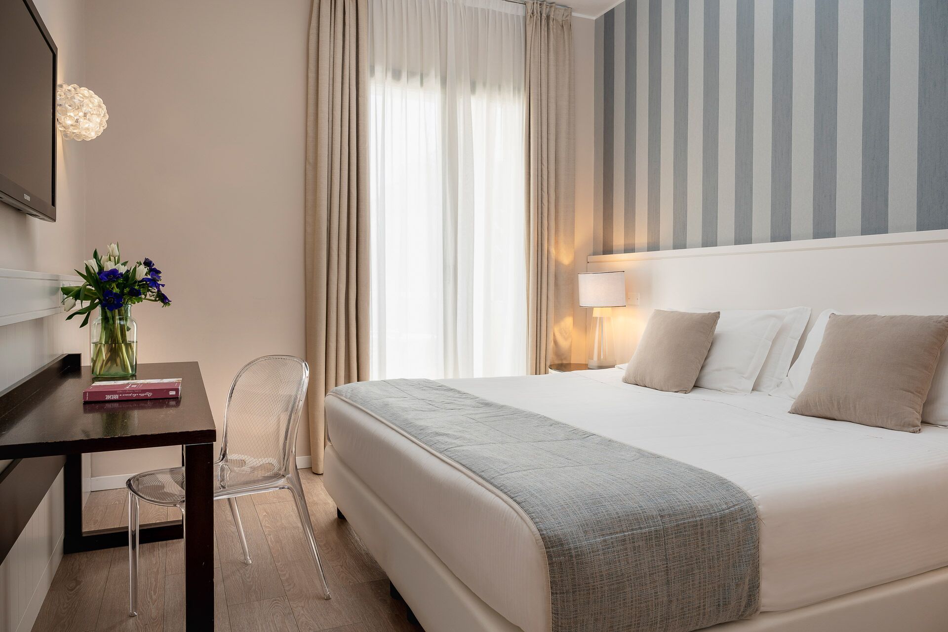 Hotellihuone, jossa on sänky, työpöytä tuolilla, maljakko kukkineen ja raidallinen seinä.