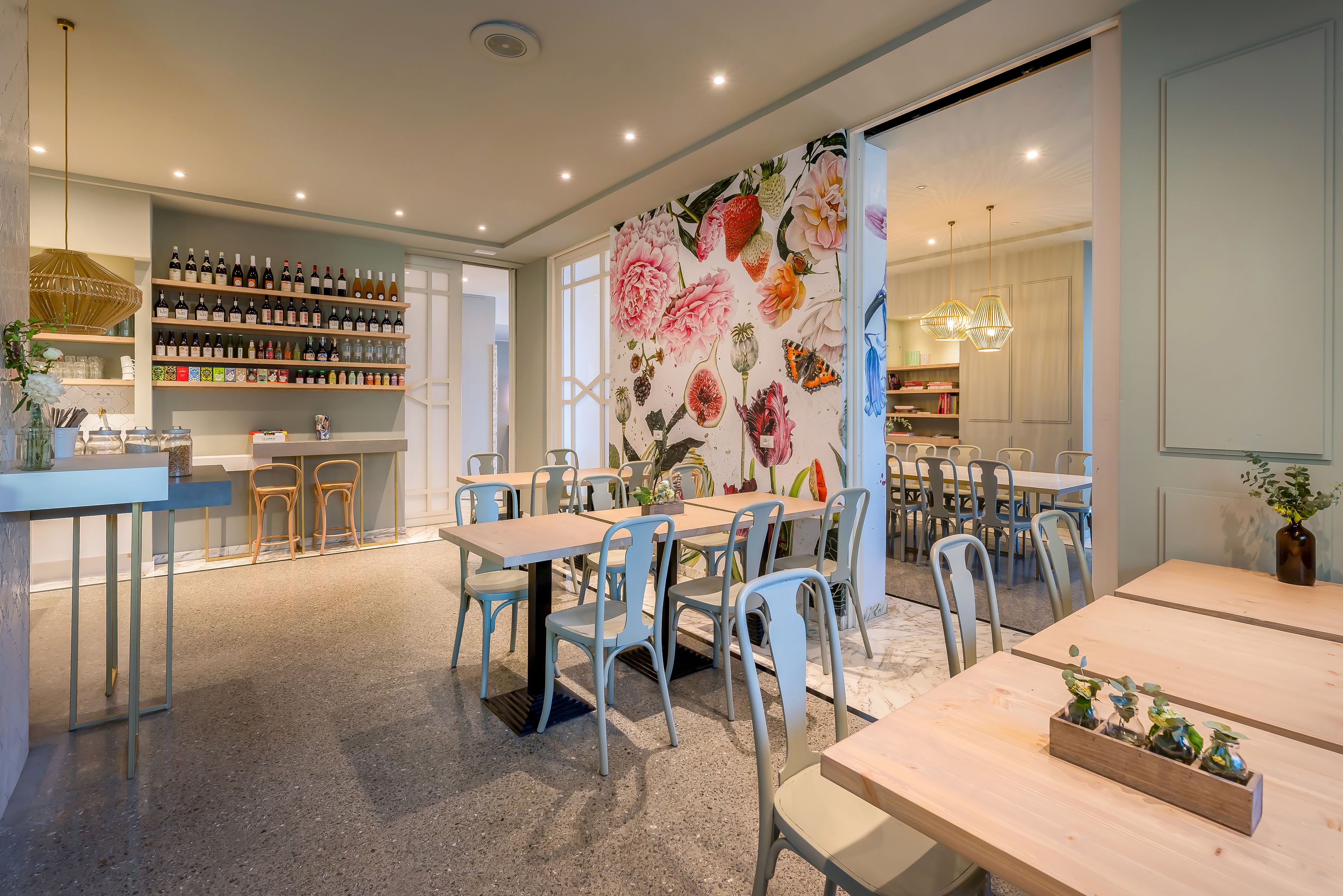 A restaurant with multiple tables and chairs, a wall with a floral mural, and shelves stocked with bottles and glasses.