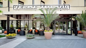 Entrada de un hotel con un letrero que dice "TERME ITALIANO". Hay áreas de estar al aire libre con sillas y mesas, además de grandes macetas con plantas.