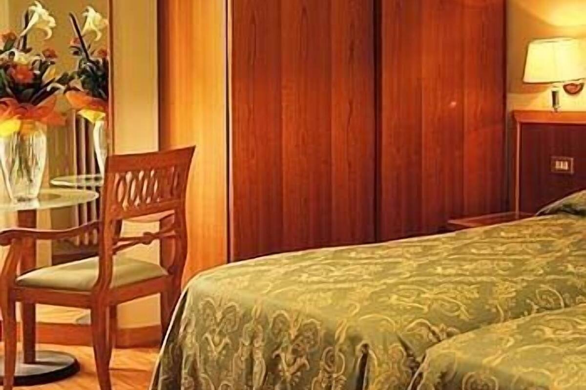 Habitación de hotel con suelo de madera, colcha estampada verde, una silla de madera, una mesita con un jarrón de flores y una mesita de noche con lámpara.