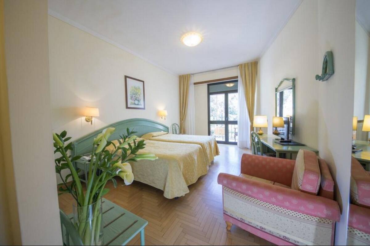 Habitación de hotel con dos camas, un área de estar con un sofá rosa, una mesa de comedor y un gran ventanal con cortinas.