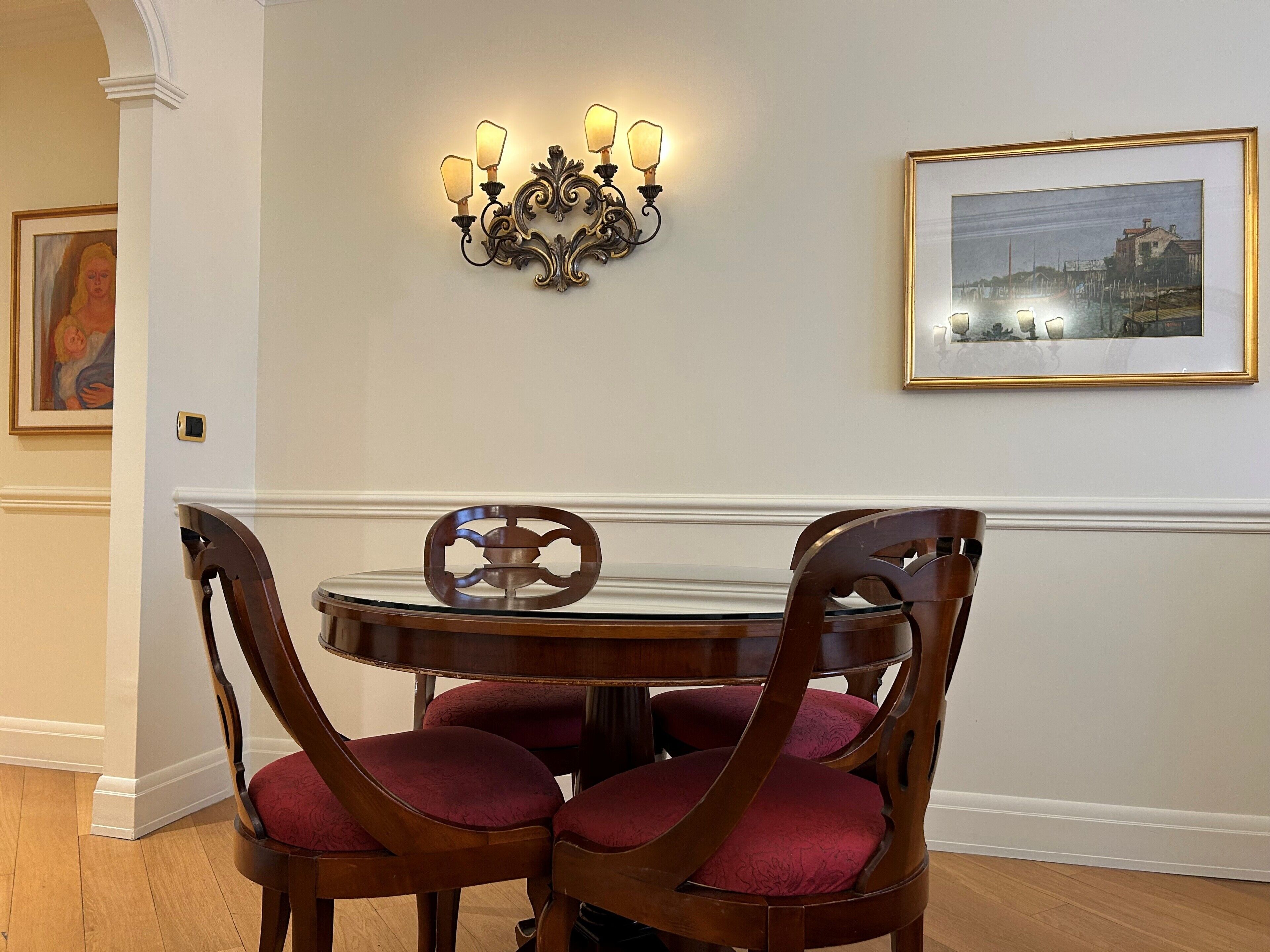 Un coin repas avec une table ronde, quatre chaises en bois dotées de coussins rouges, un luminaire fixé au mur et un tableau encadré.