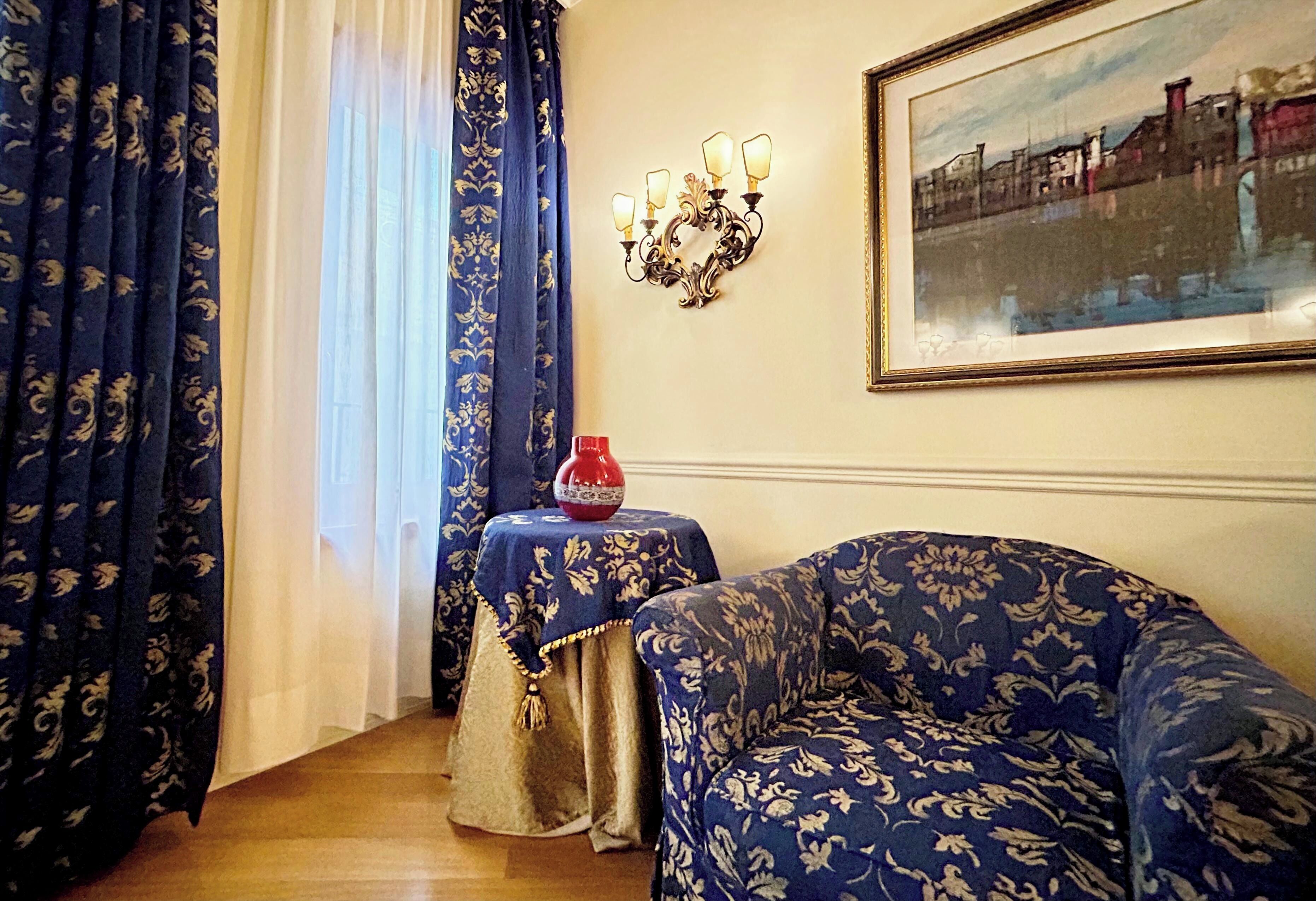 Una habitación con un sofá estampado, una mesita con un florero, una lámpara de pared e imagen enmarcadas.