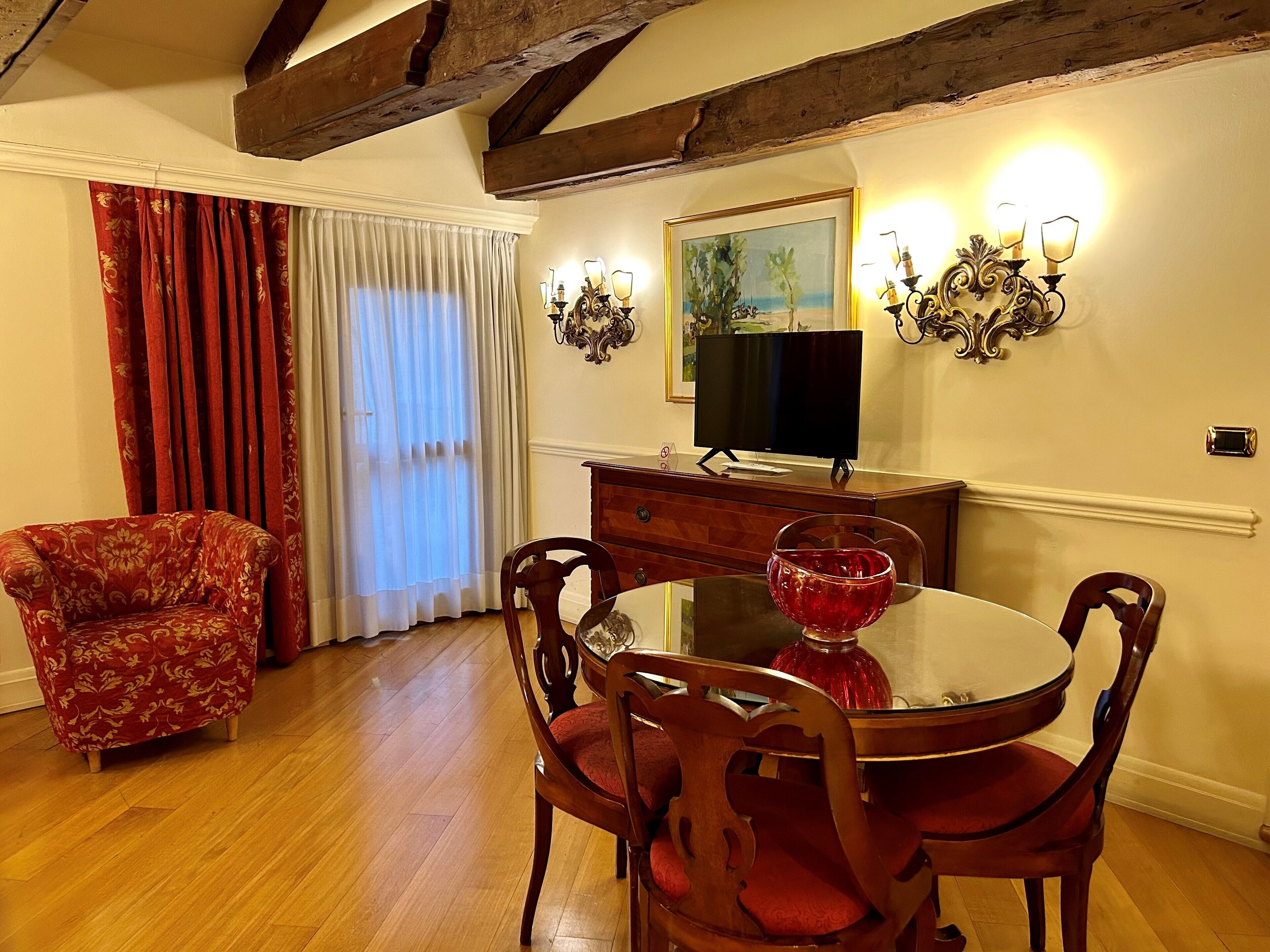 Ein Essbereich mit Holztisch, rot gepolsterten Stühlen, einer roten Glasvase, einem Fernseher auf einer Holzkommode und einem Bild an der Wand.
