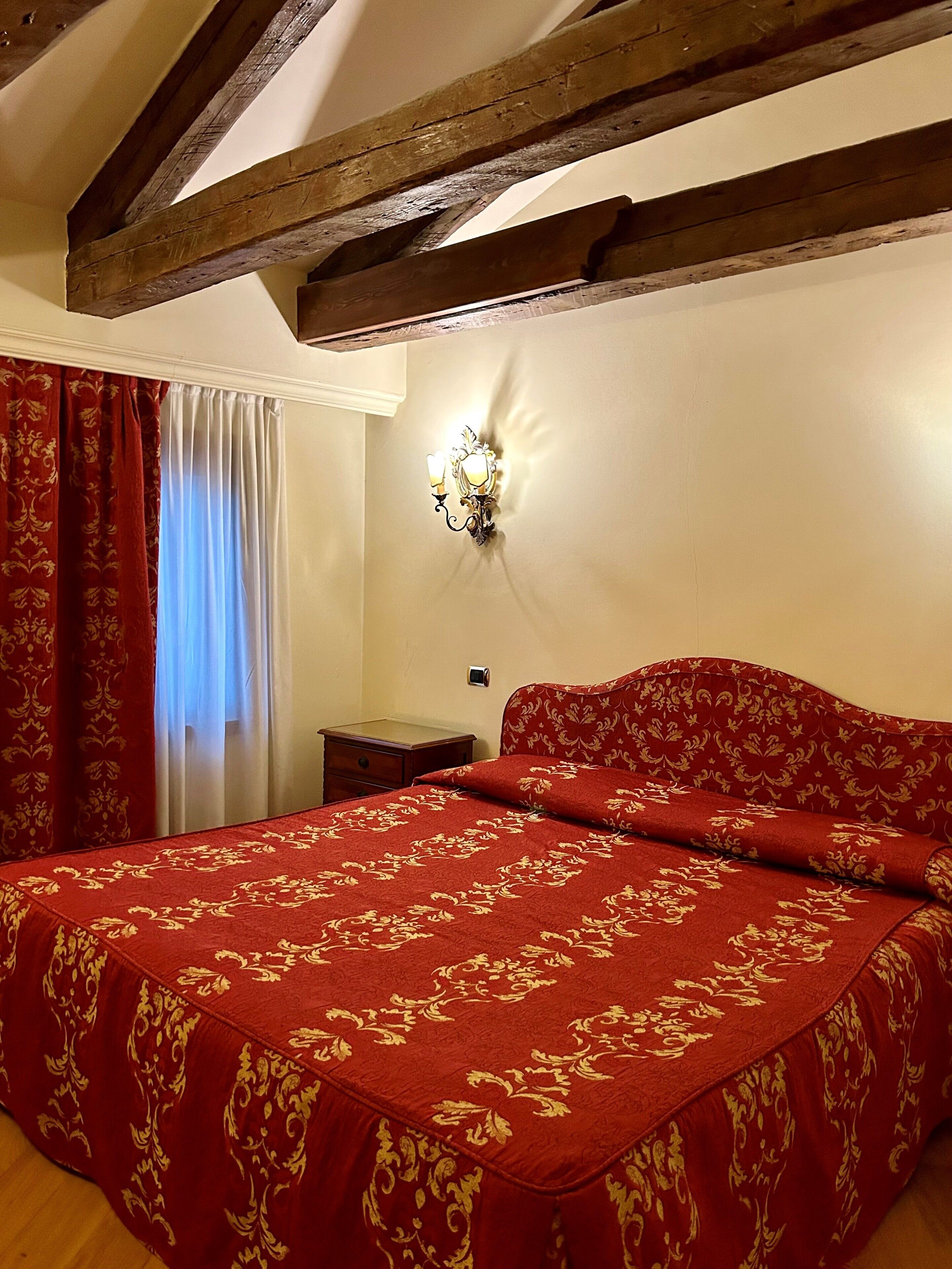 A bedroom with a large bed, wooden beams, a wall-mounted light fixture, and a window with curtains.