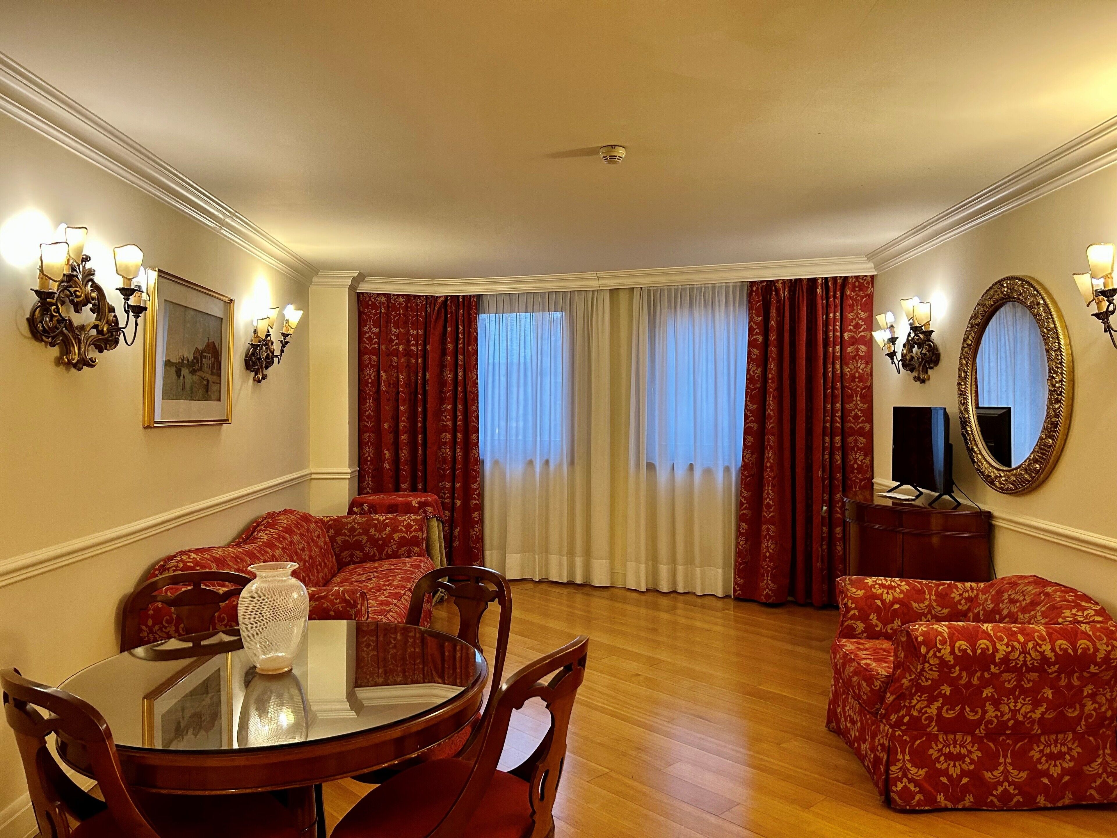 Uma sala de estar espaçosa com uma mesa de jantar redonda, sofás vermelhos estampados, um espelho grande e uma televisão em um armário de madeira.
