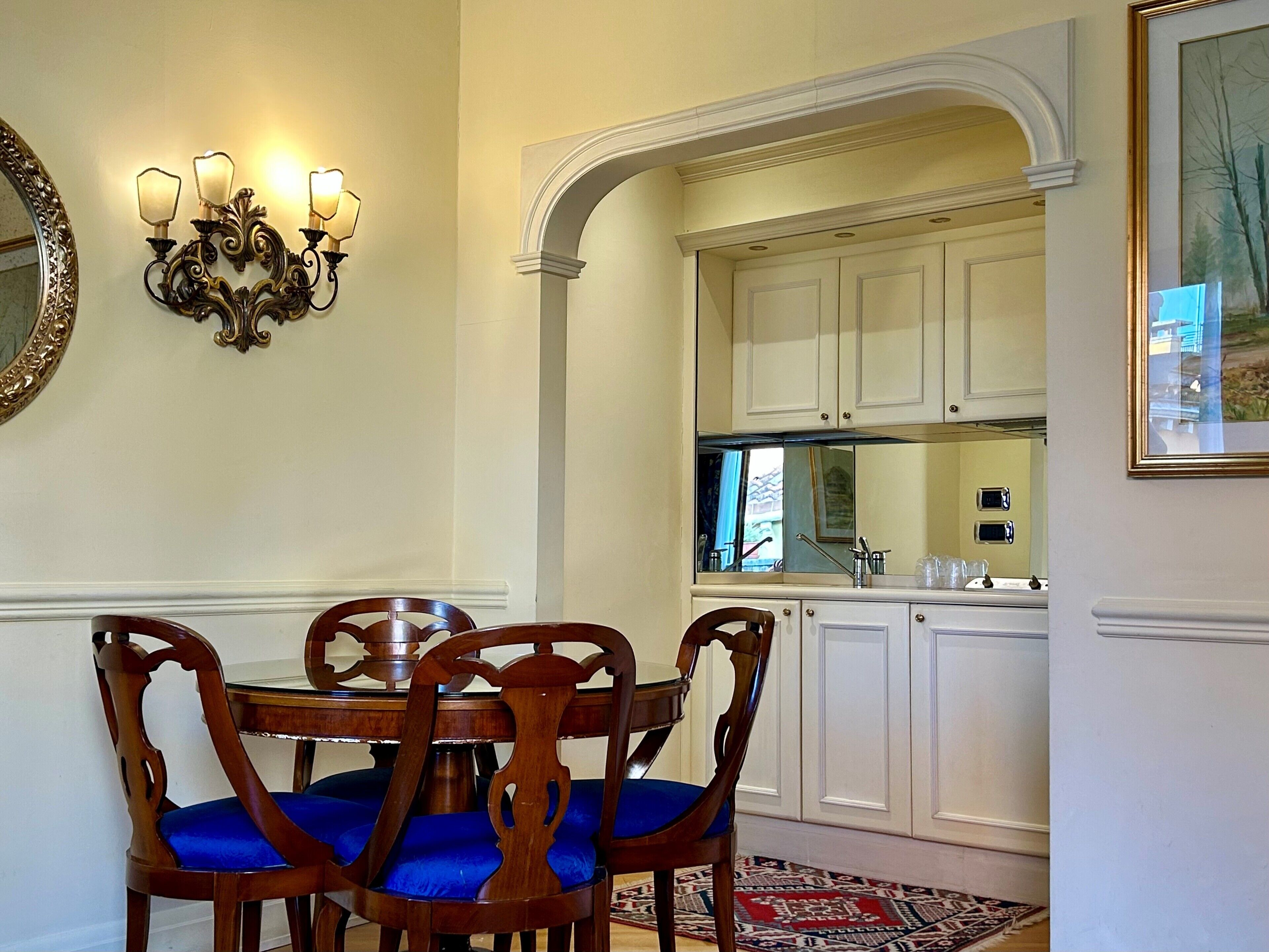A dining area with a round table and four chairs, a wall-mounted light fixture, and a kitchen visible through an archway.