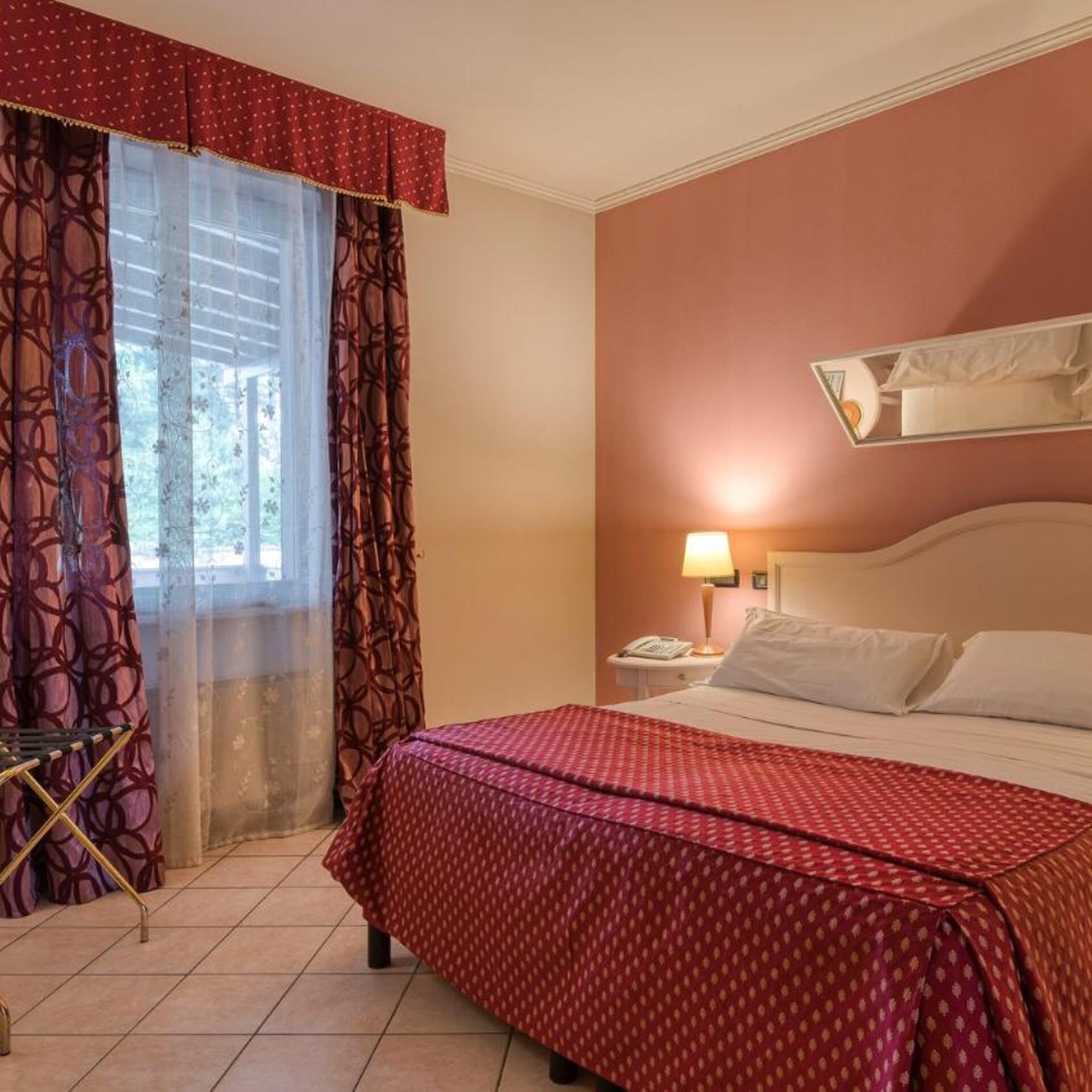 A hotel room with a bed, a nightstand with a lamp, a window with curtains, and a wall-mounted light fixture.