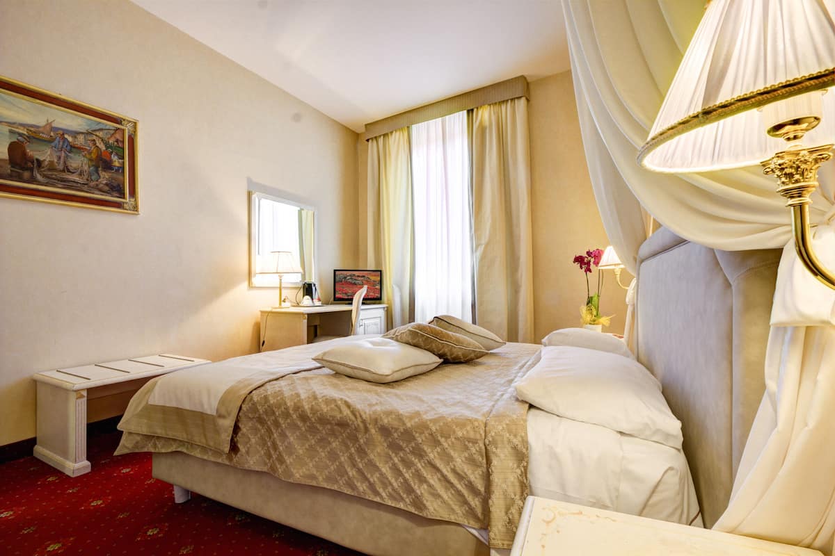 A hotel room with a large bed, a desk with a computer, a window with curtains, and a painting on the wall.