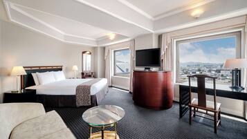 A hotel room with a bed, a TV, a desk, a chair, and a view of the city.