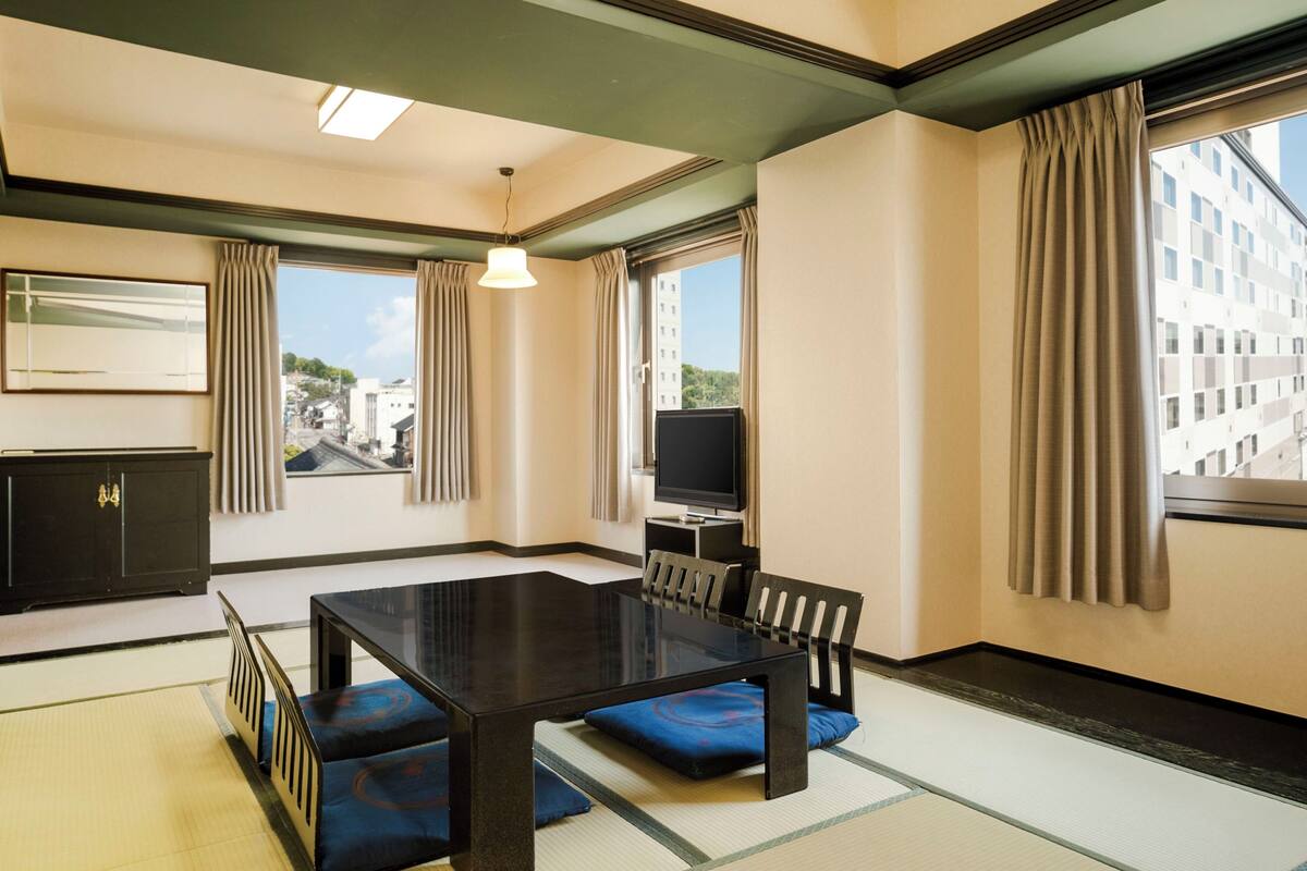 Une chambre d’hôtel avec un sol recouvert de tatamis, une table à manger, des chaises, un téléviseur et de grandes fenêtres avec des rideaux.