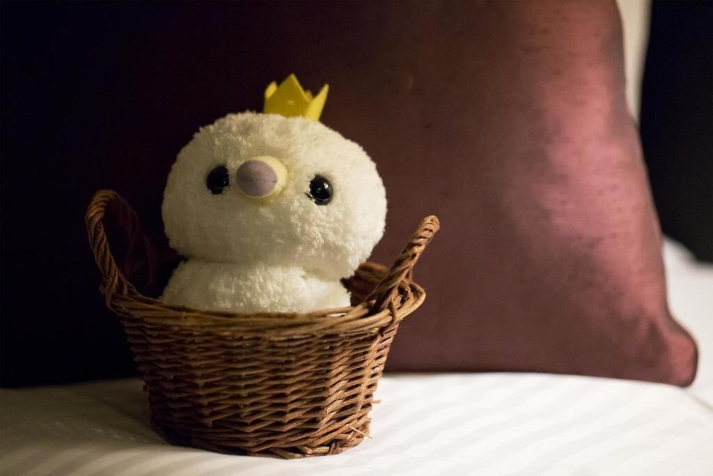 A plush toy in a wicker basket on a bed with a dark pillow in the background.