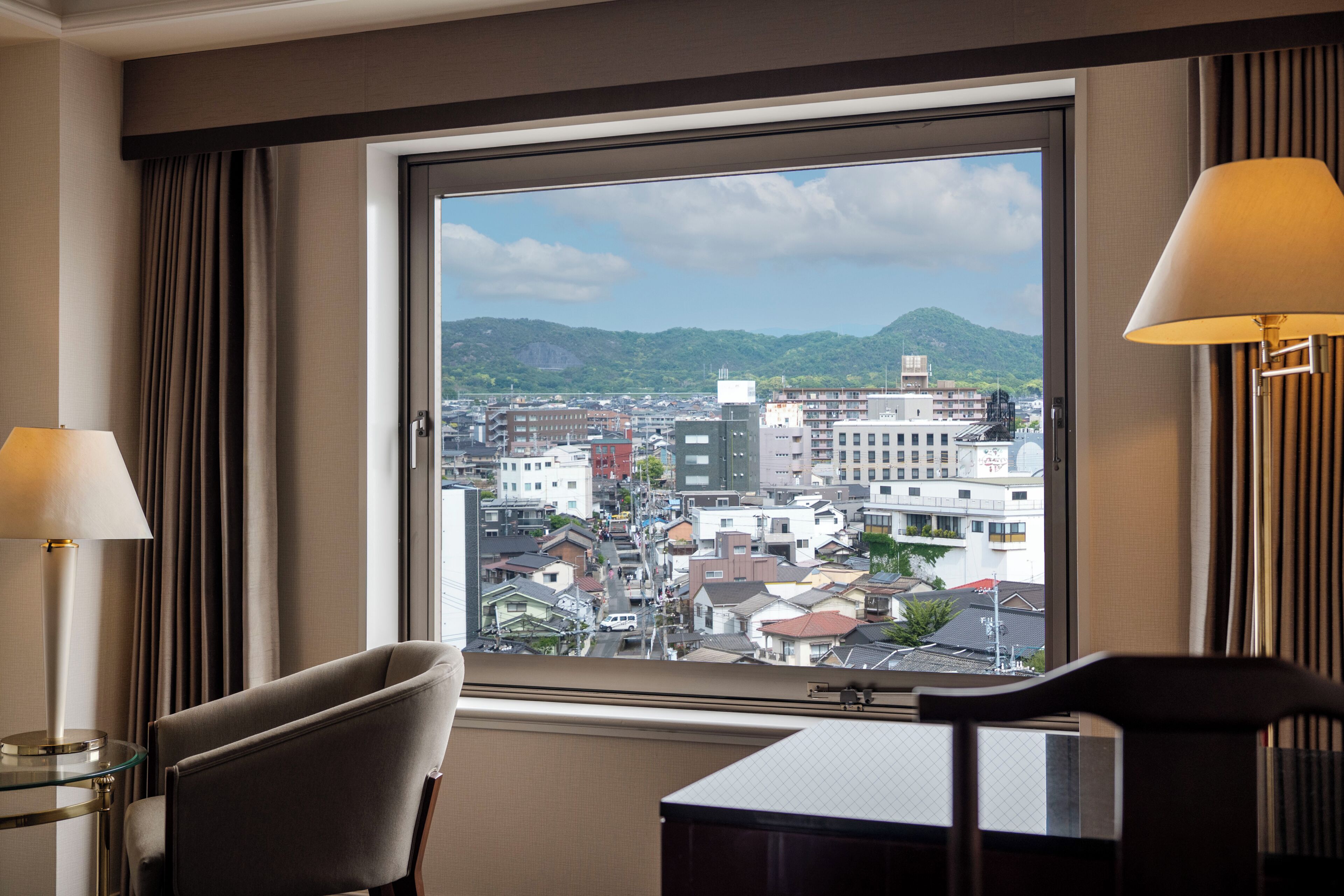A hotel room with a large window, a chair, a table, and two lamps.