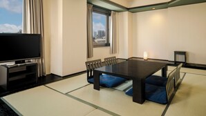 Une chambre d’hôtel avec un sol recouvert de tatamis, une grande tél évision à écran plat, une table à manger avec des chaises et une vue sur la ville.