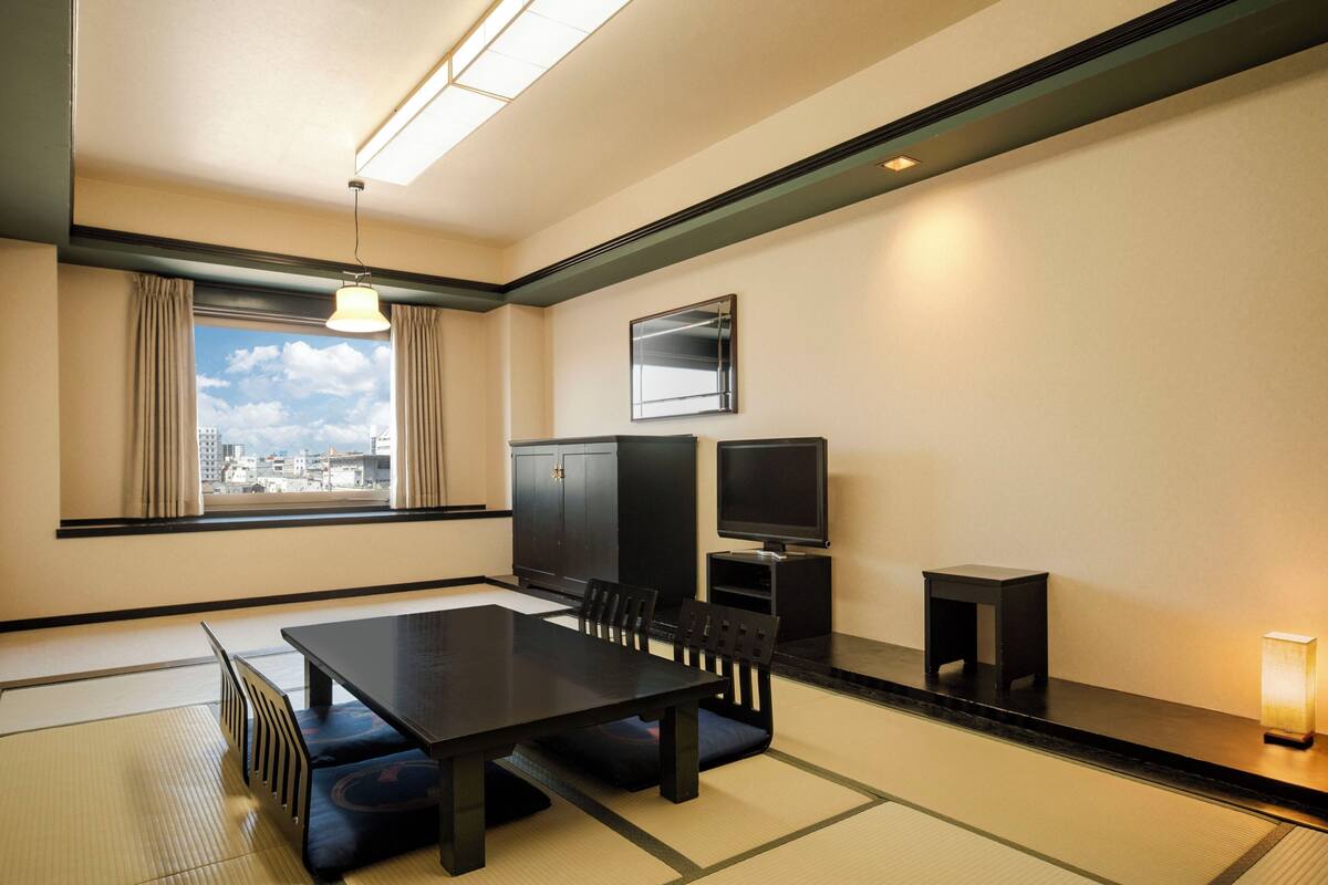Une chambre d’hôtel avec un sol recouvert de tatamis, une table à manger avec des chaises, un téléviseur, une armoire et une fenêtre avec des rideaux.