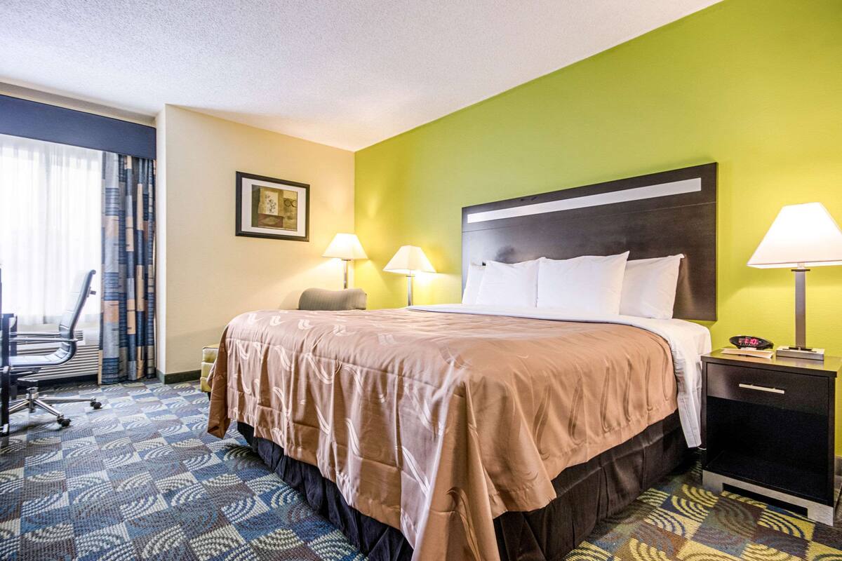 A hotel room with a bed, two bedside lamps, a desk with a chair, and a patterned carpet.