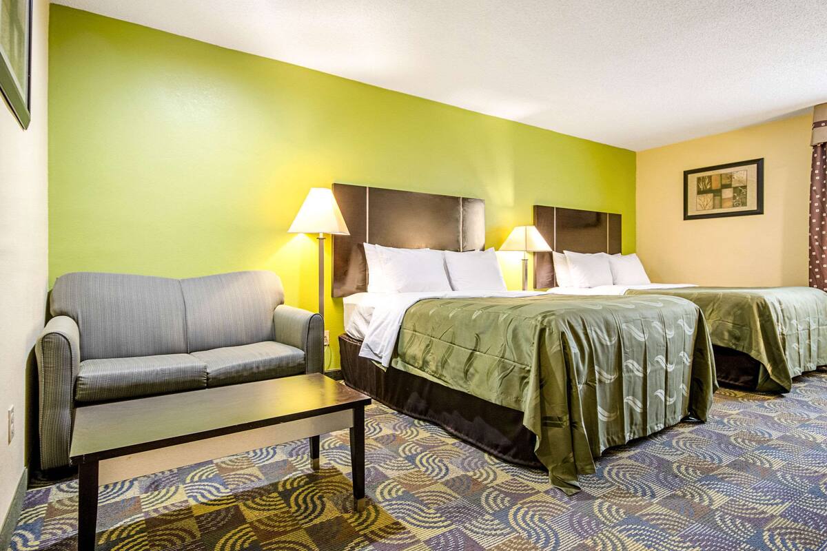 A hotel room with a green wall, two beds with green bedspreads, a grey sofa, and a small table.
