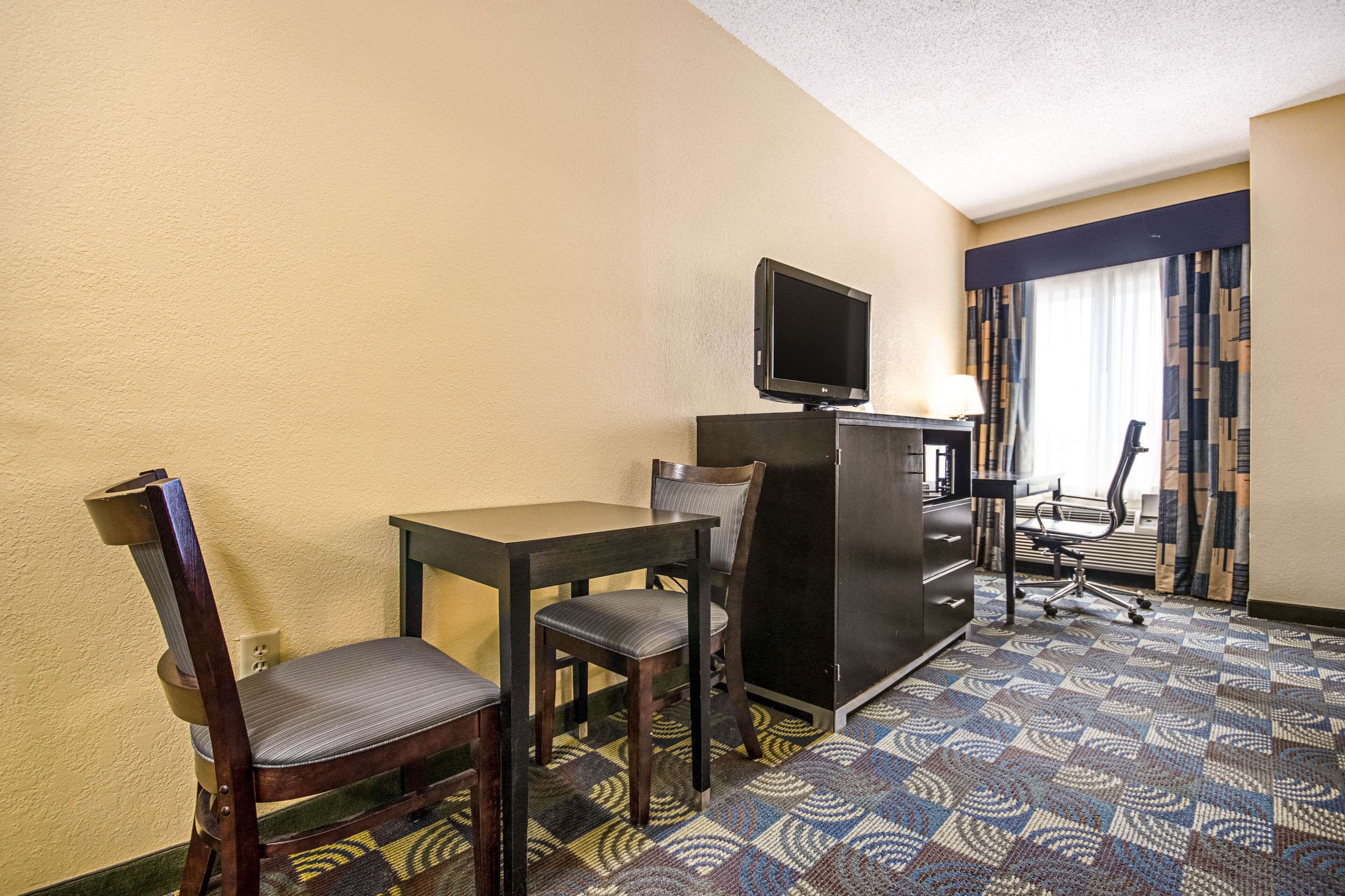 A hotel room with a desk, two chairs, a television, a desk lamp, and a patterned carpet.