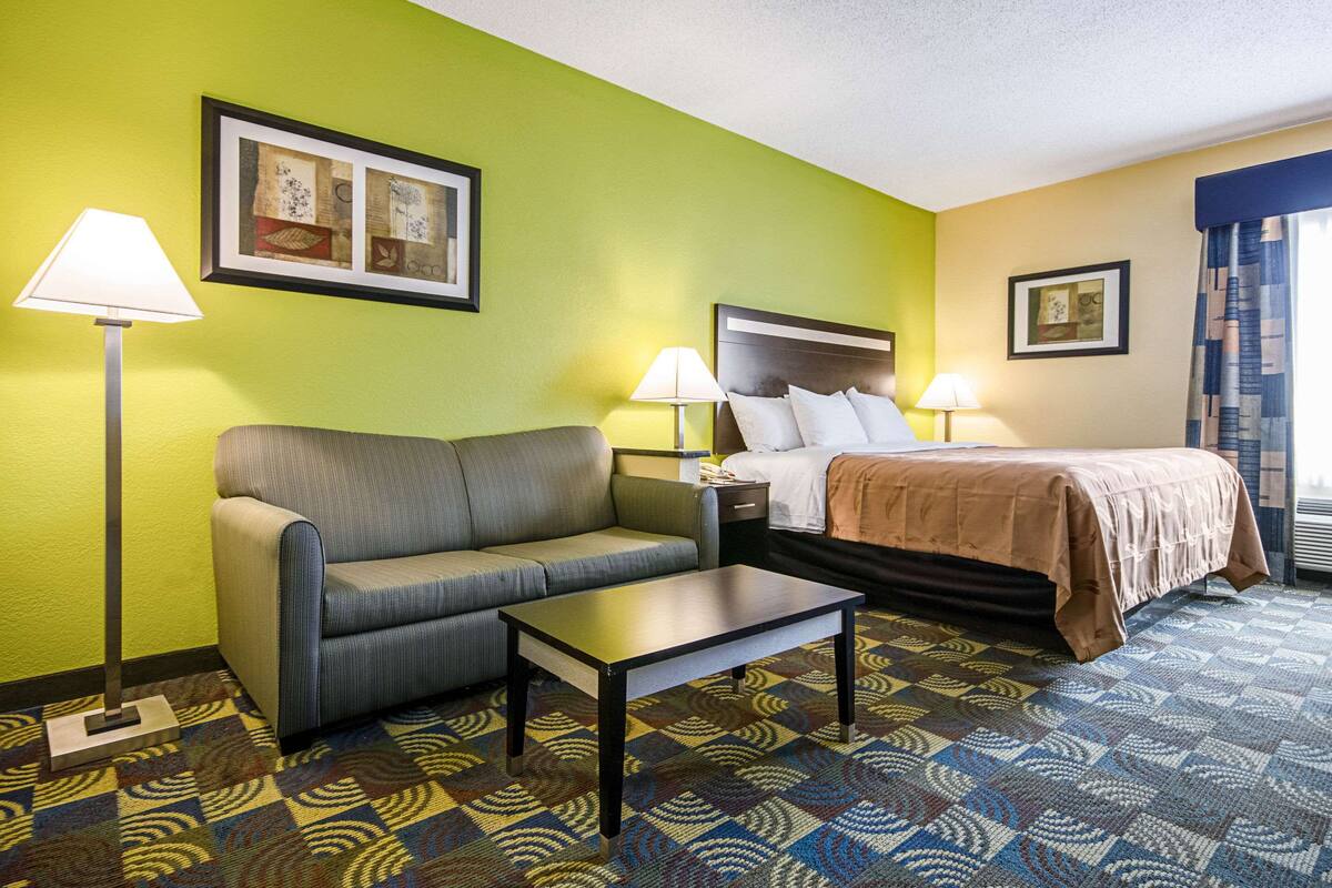 A hotel room with a green wall, a bed with a brown blanket, a gray sofa, a small table, and two lamps.