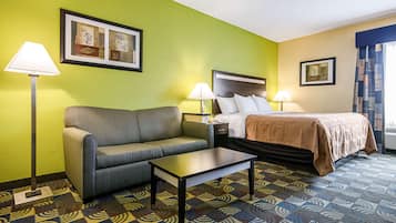 Une chambre d’hôtel avec un mur vert, un lit recouvert d’une couverture marron, un canapé gris, une petite table et deux lampes.