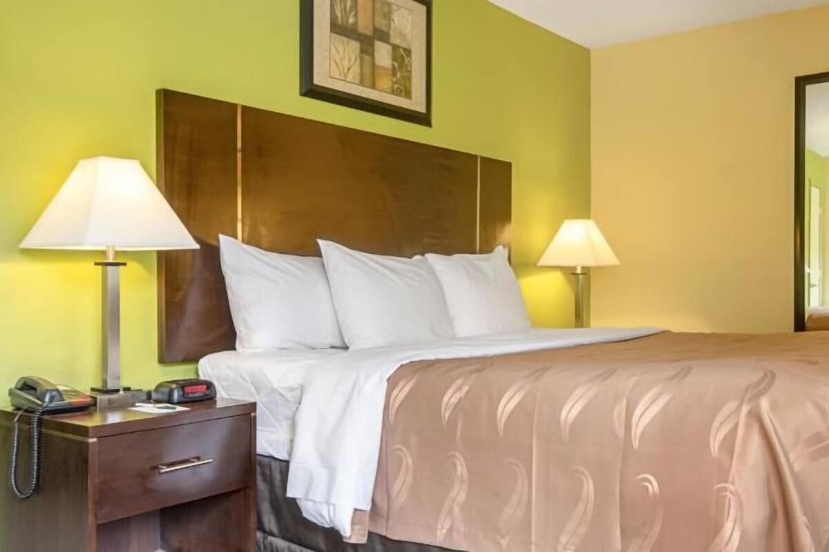 A hotel room with a bed, two bedside lamps, a telephone, a desk with a lamp, and a mirror.