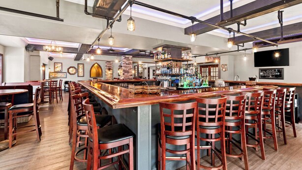 A bar area with wooden bar stools, a long counter, and hanging light fixtures. - Genetti Hotel, SureStay Collection by Best Western (Williamsport)