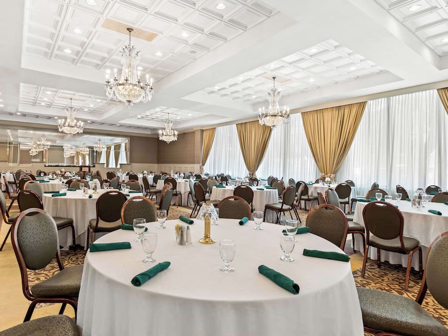 A banquet hall with round tables, chairs, chandeliers, and large windows with curtains.
