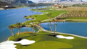 Egy golfpálya, melyet egy víztér, pálmafák és hegyek alkotják a háttérben.