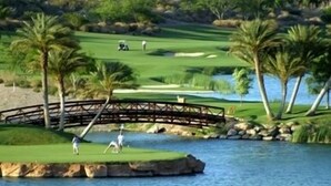Egy golfpálya, amelyen híd ível át a víz felett, pálmafák állnak, és a felhőtlen égbolt látható.