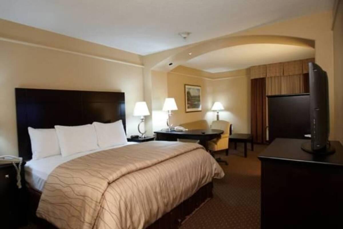 A hotel room with a large bed, two bedside tables with lamps, a desk, a chair, a television, and a small kitchenette area.