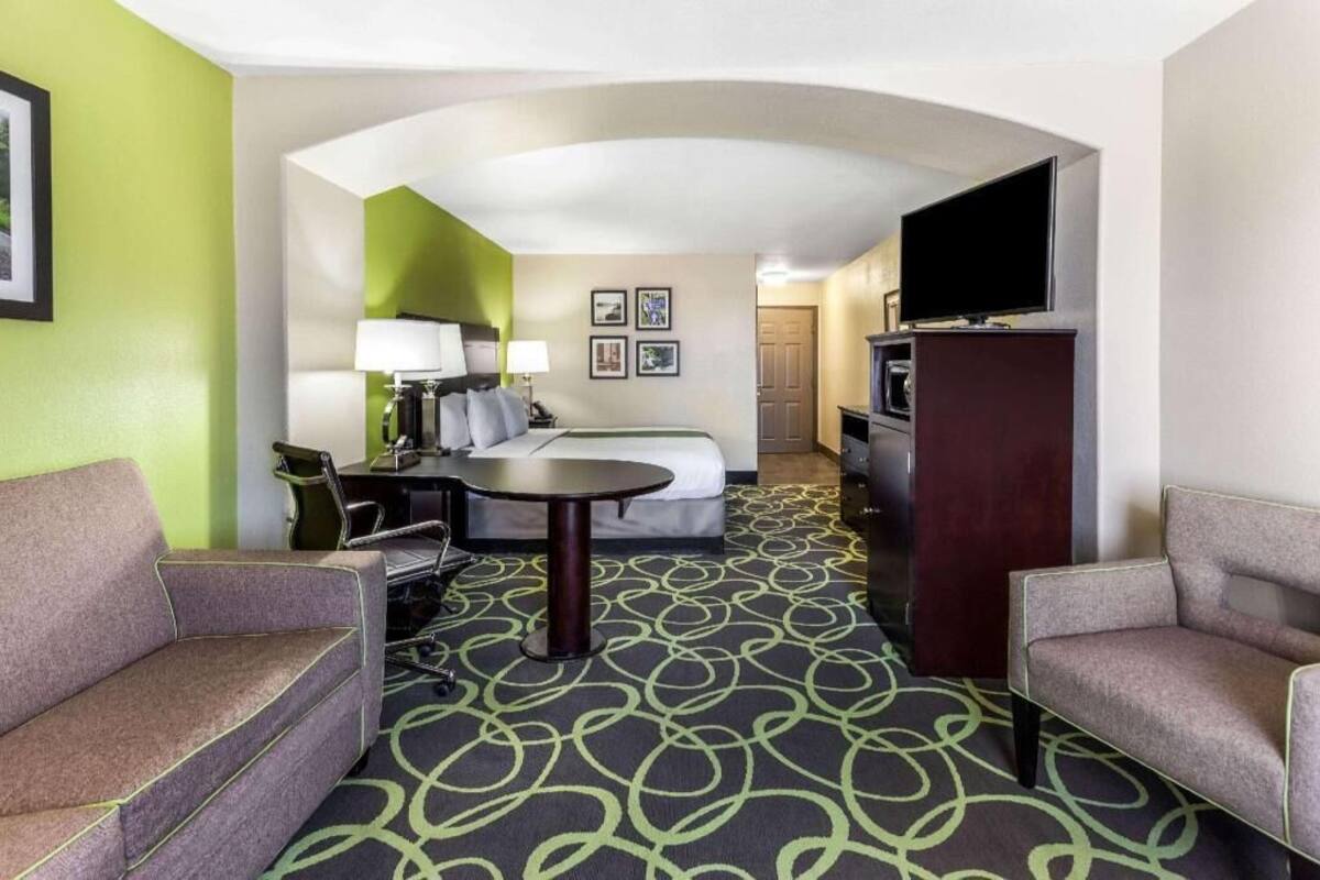 A hotel room with a bed, two armchairs, a small table, a TV, and a desk with a lamp.