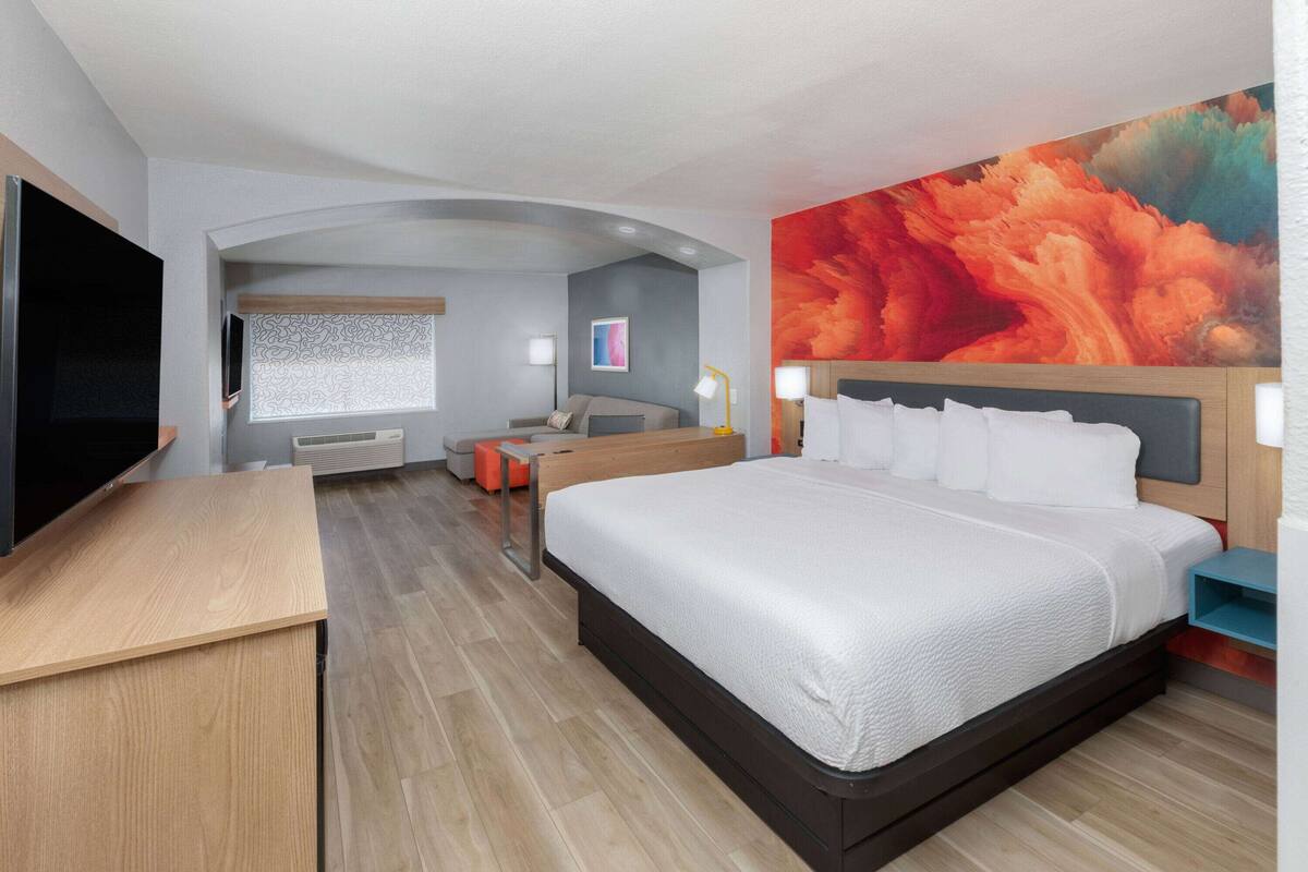 A modern hotel room with a large bed, a desk, a TV, and a colorful mural on the wall.