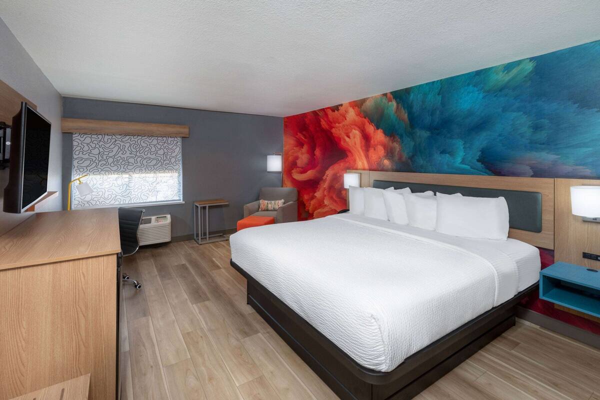 A modern hotel room with a large bed, a desk, a chair, a flat-screen TV, and a colorful mural on the wall.
