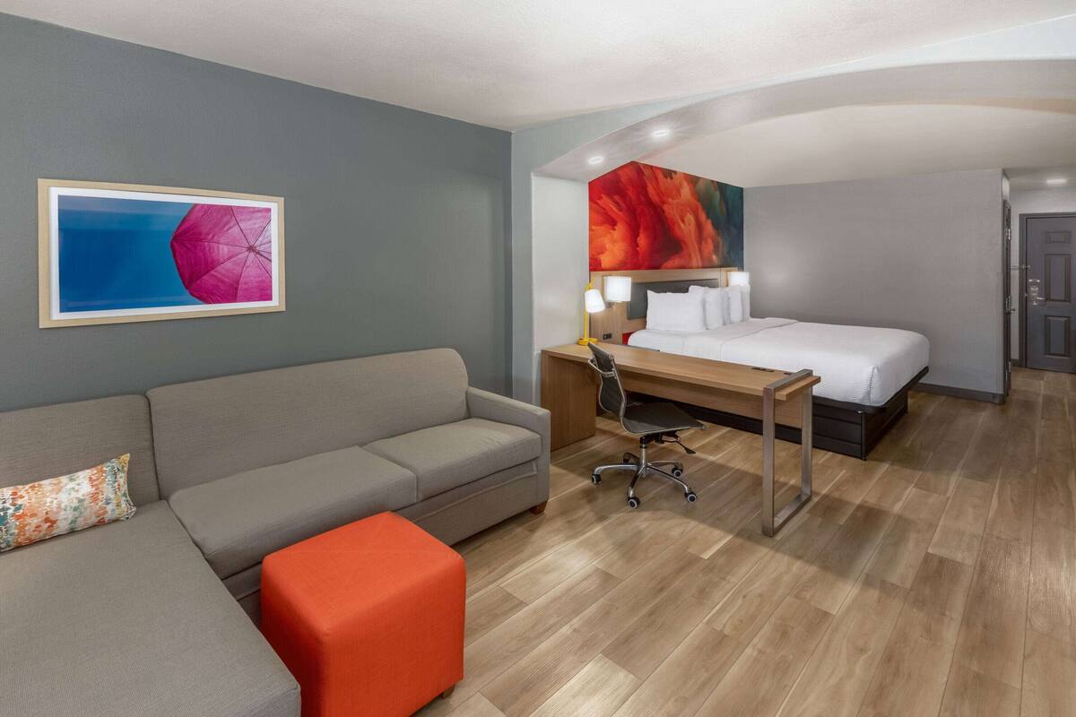 A hotel room with a grey sofa, a wooden desk, a bed with white linens, and a colorful abstract painting on the wall.
