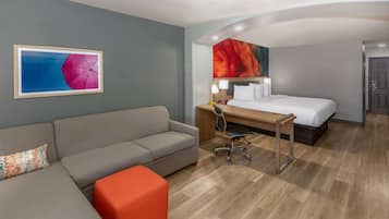 Habitación de hotel con sofá gris, un escritorio de madera, cama con ropa blanca y un cuadro abstracto colorido en la pared.