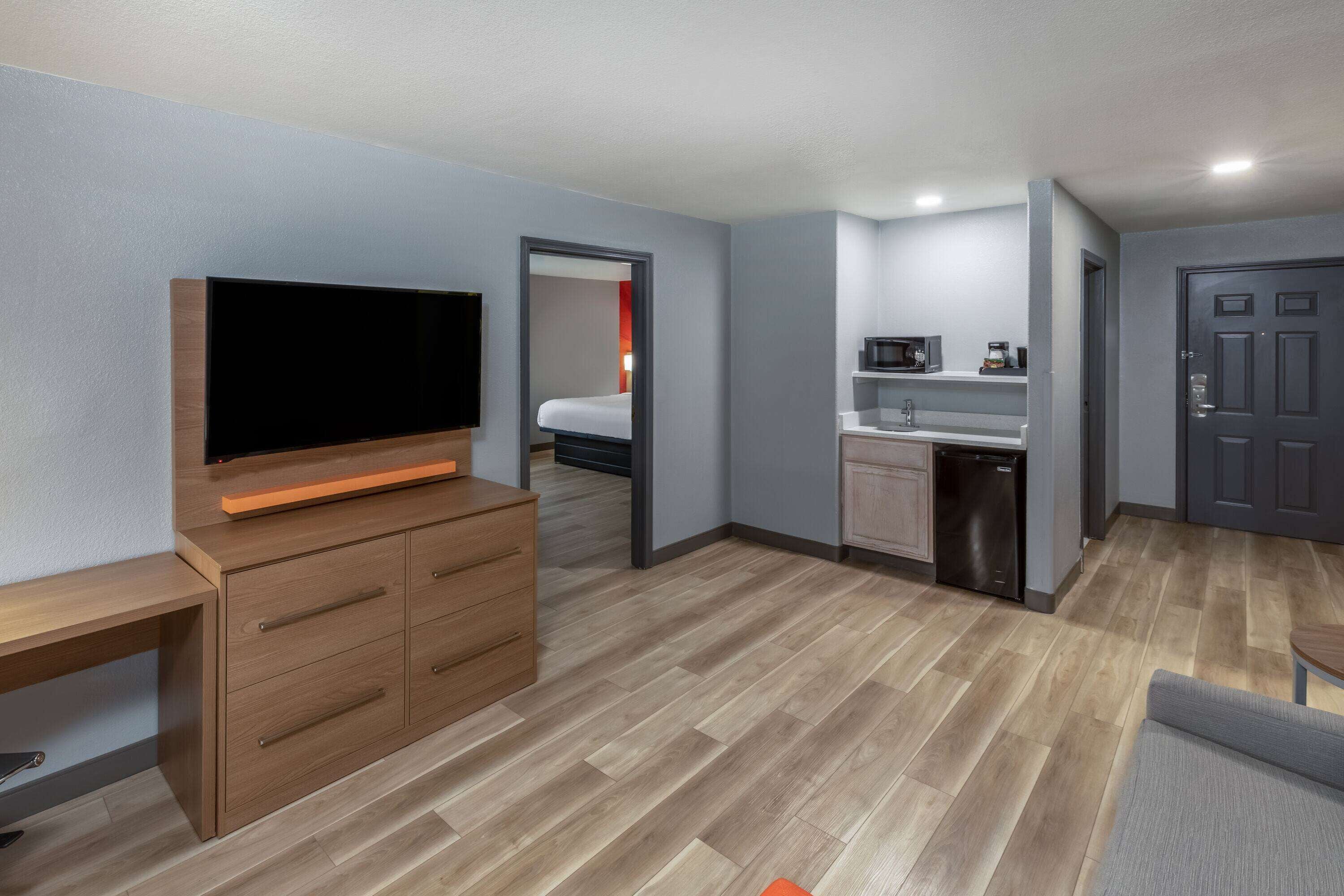 Une chambre d’hôtel de taille réduite, dotée d’une télévision, d’une commode en bois, d’une kitchenette et d’une porte.