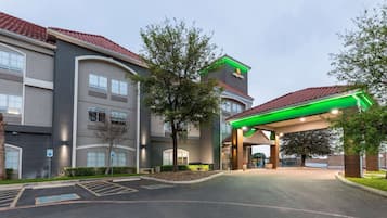 A hotel entrance with a covered driveway, a parking lot, and a building with a red-tiled roof and a green-lit sign.