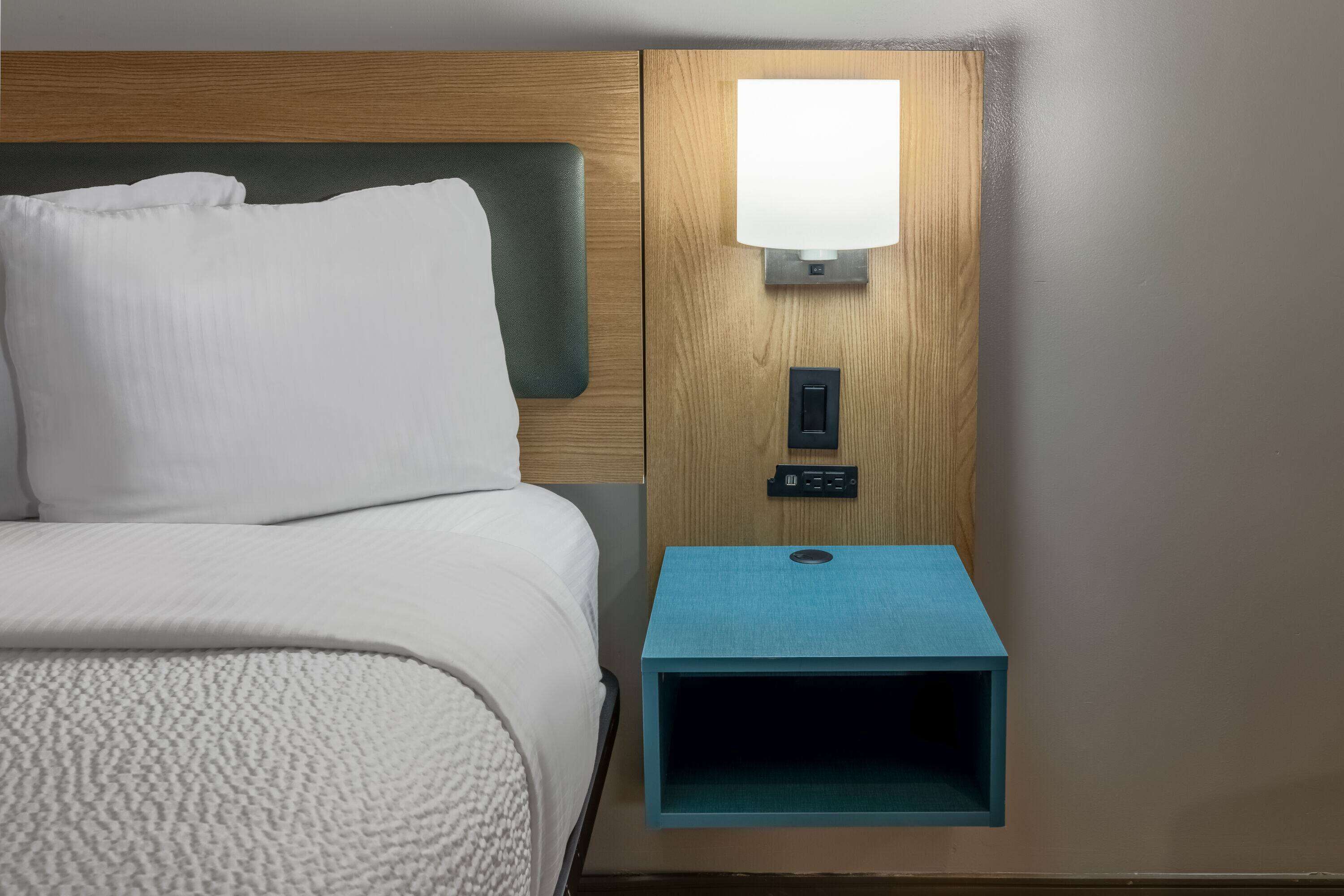 A hotel room with a neatly made bed, a wooden headboard, a nightstand with a lamp, and a small blue storage box.