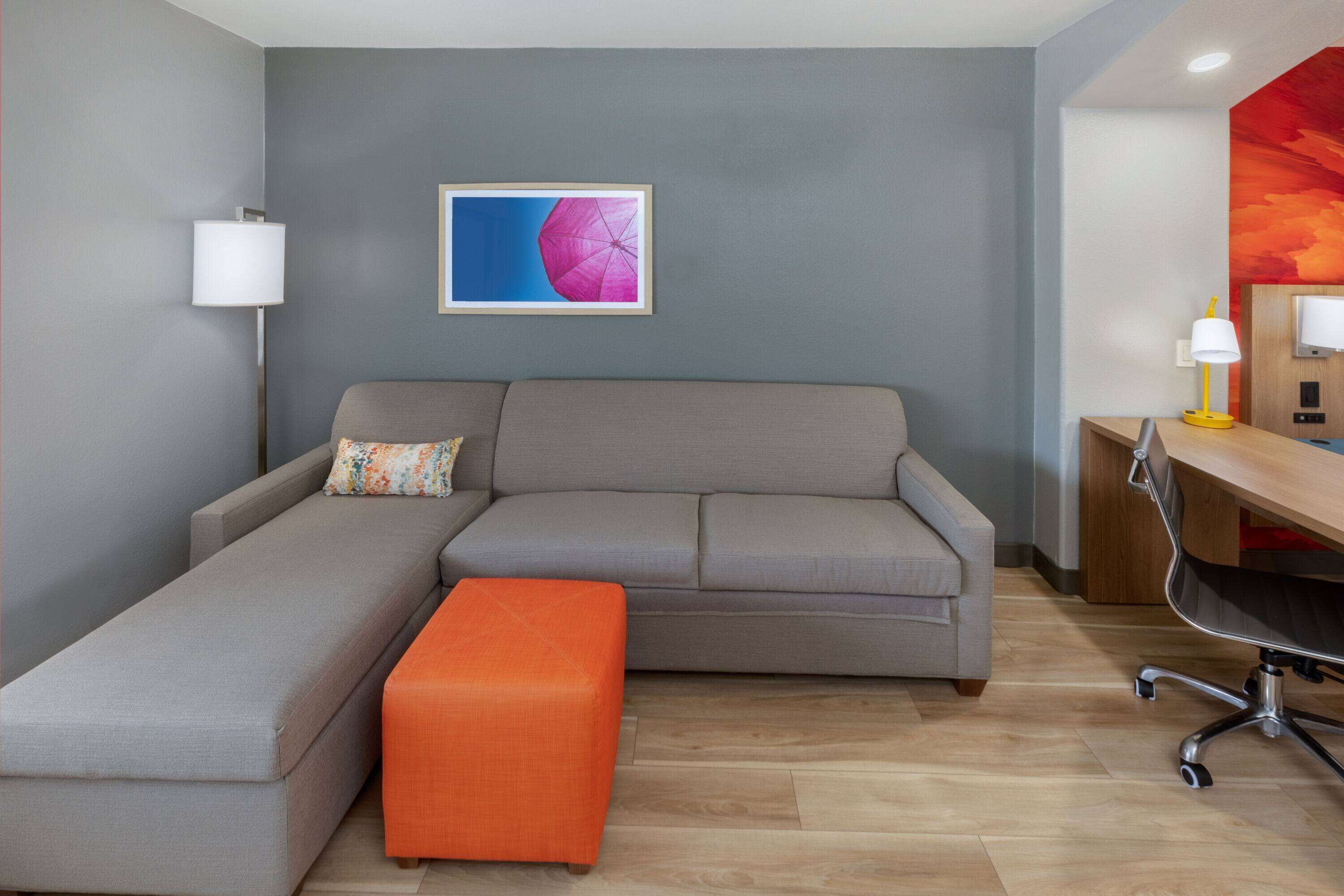 A modern living room with a grey sofa, an orange ottoman, a desk with a lamp, and a framed picture on the wall.