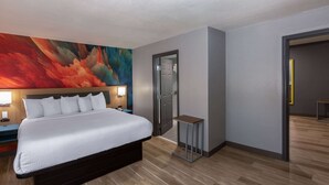 Ein modernes Hotelzimmer mit einem großen Bett, einem Nachttisch, einem kleinen Tisch und einem farbenfrohen, abstrakten Gemälde an der Wand.