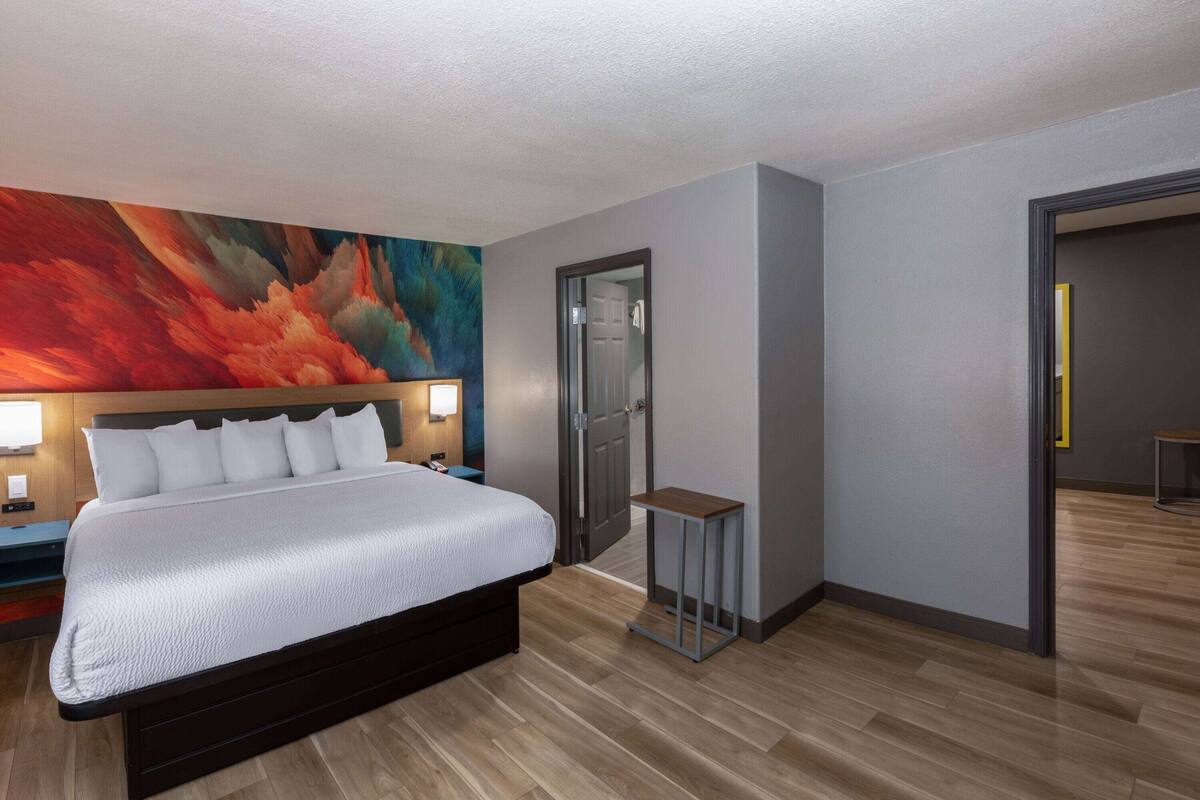 A modern hotel room with a large bed, a nightstand, a small table, and a vibrant abstract painting on the wall.