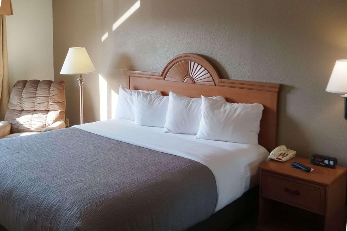 A hotel room with a bed, a nightstand with a phone and remote, a chair, and a lamp.