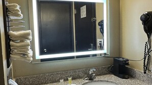 Un baño con encimera de granito, lavabo, espejo con luz, secador de pelo y toallas en un perchero.