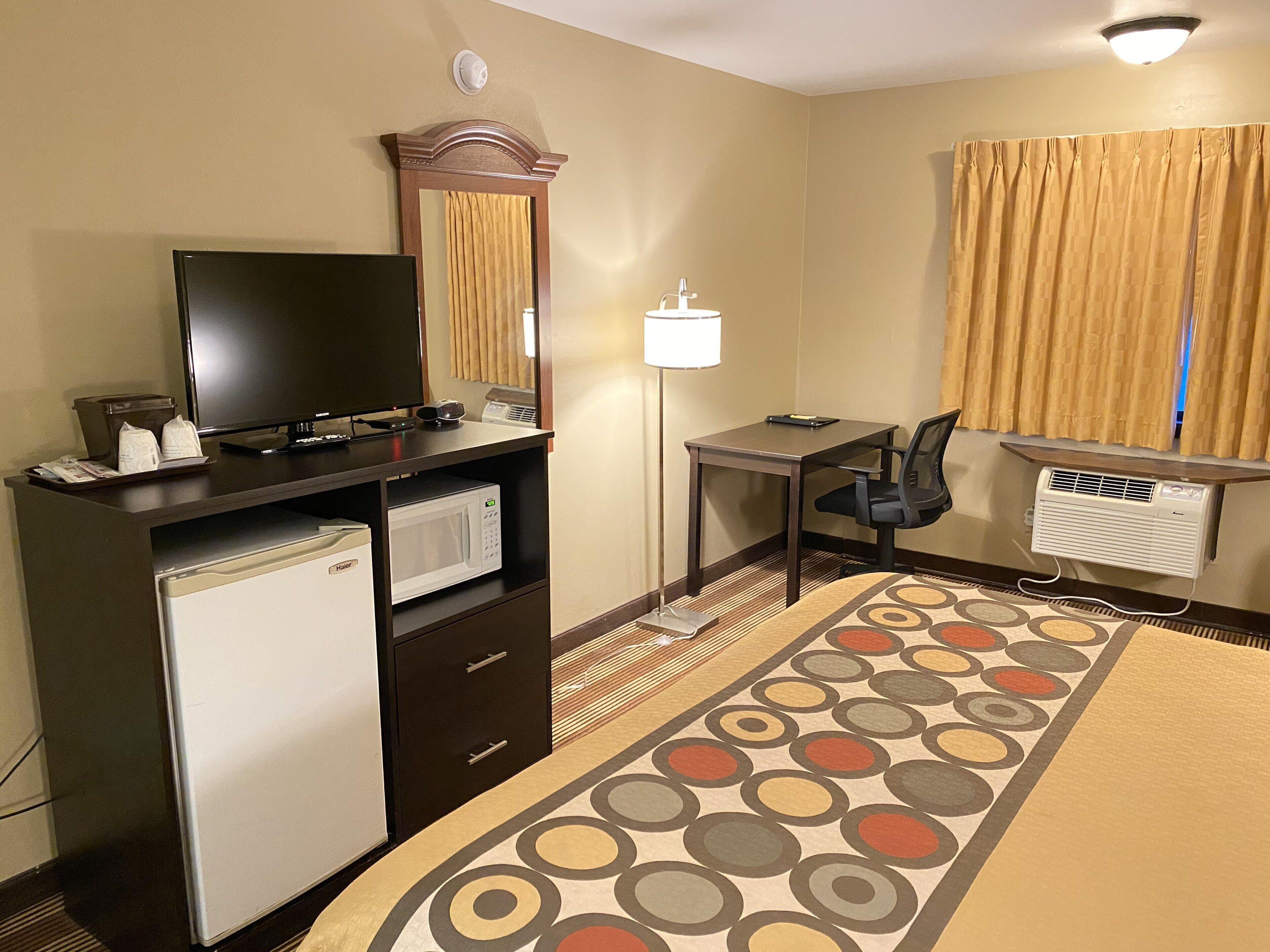 Habitación de hotel con escritorio, silla, televisor, mini refrigerador y una alfombra estampada.